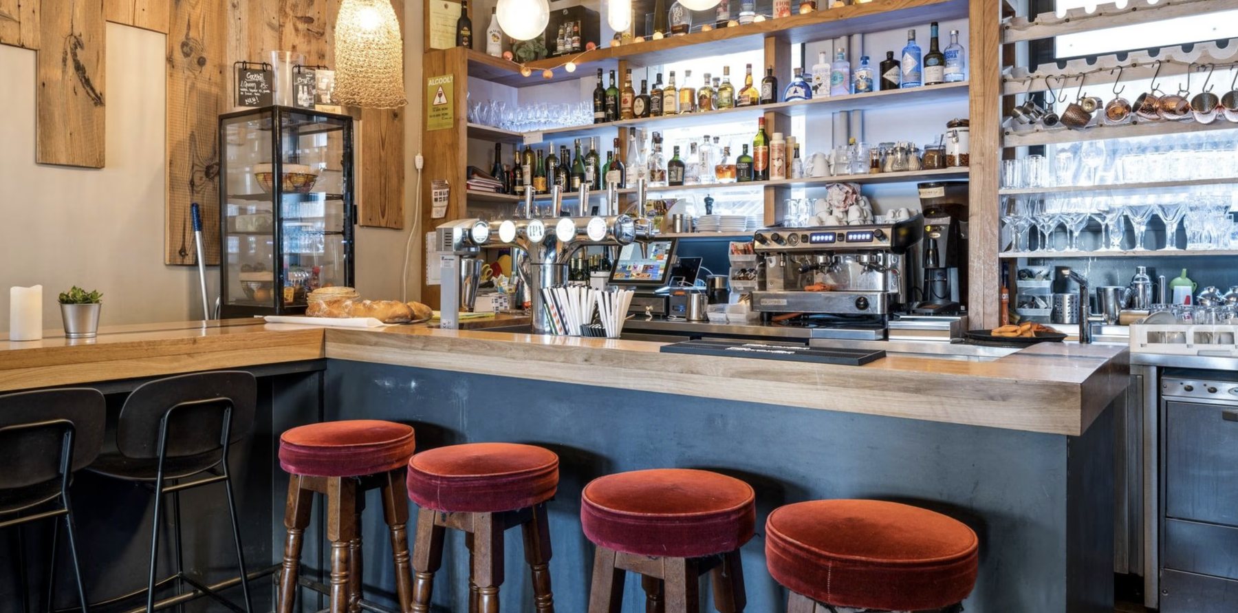 Le bar de L’Épi Doré, comptoir en bois, tabourets velours et étagères de verres.